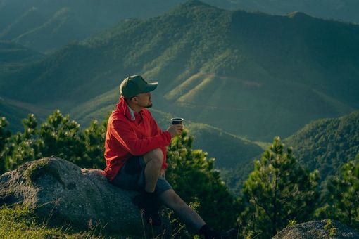 Person in a red jacket and green cap sitting on a rock, overlooking a mountain valley.