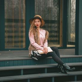 Person in a pink shirt and hat sits on steps outside a teal storefront.