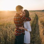 Couple embracing in a sunlit field at sunset, with tall grass and a warm golden sky.
