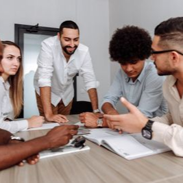Team meeting around a conference table, with one person standing and others discussing documents.