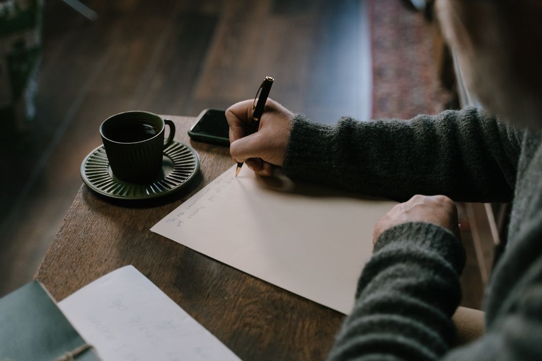 Person writing in a notebook beside a coffee cup on a wooden table