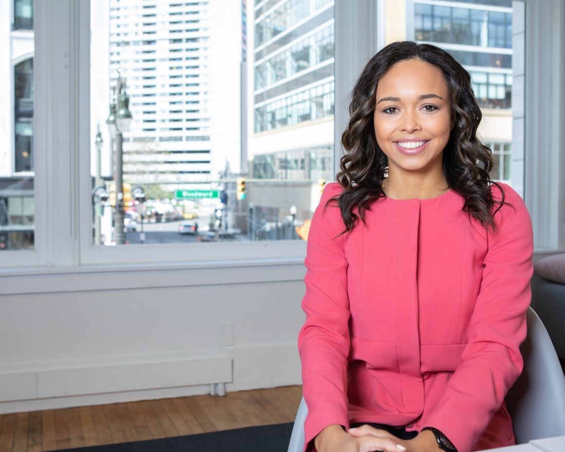 Woman in a pink blouse smiling in an office with a city view through large windows