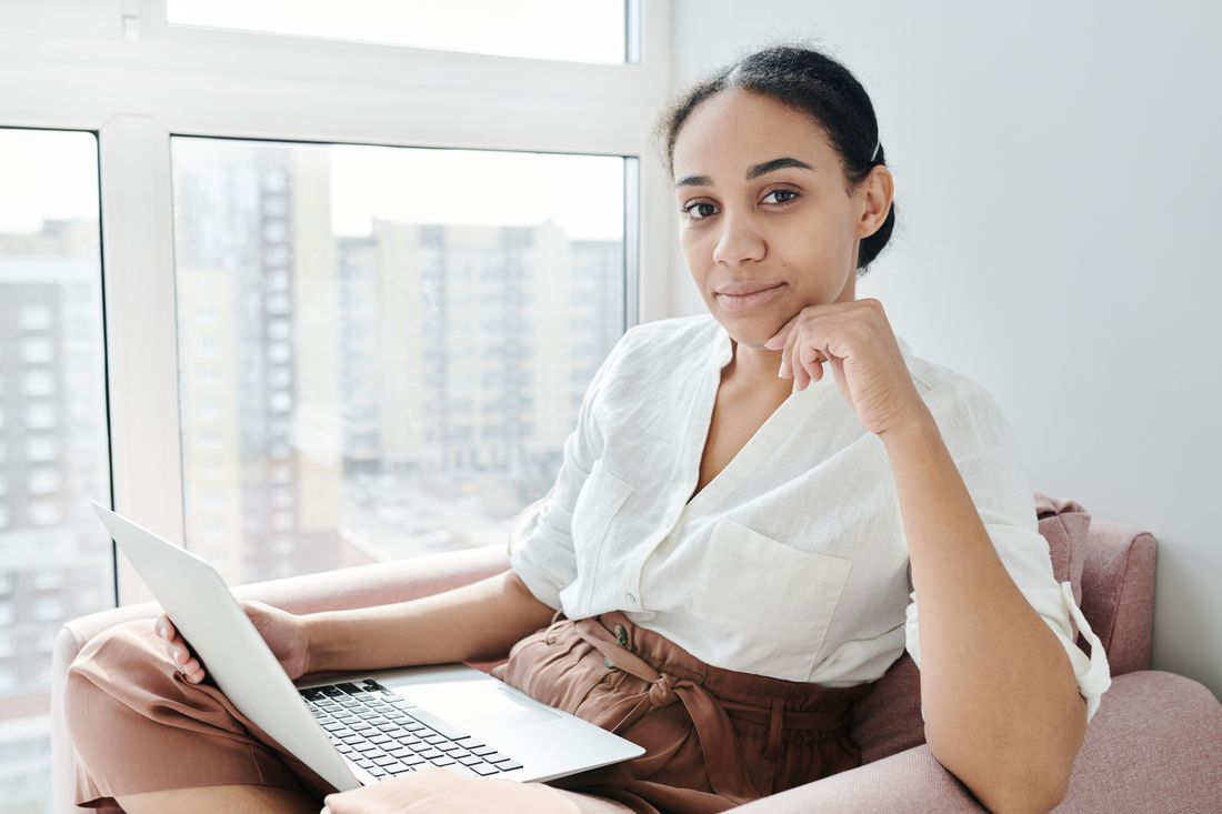 Woman using a laptop on a couch in a bright room, looking at the camera.