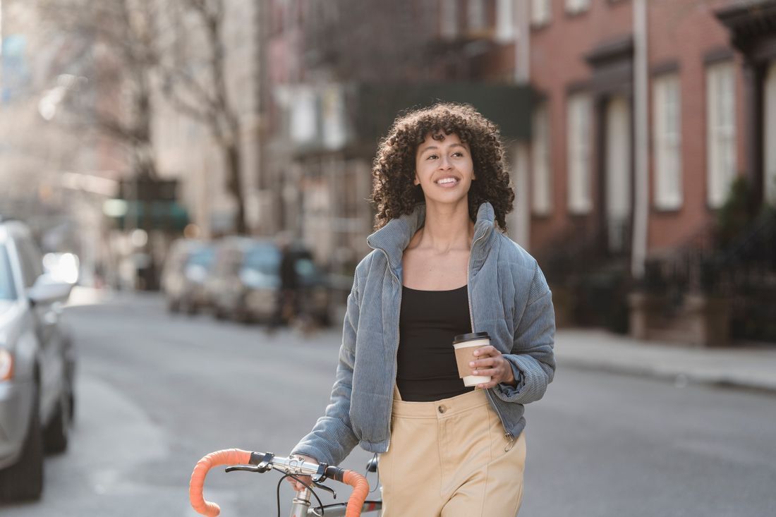Smiling person walking a bicycle on a city street, holding a drink and wearing a denim shirt and tan pants