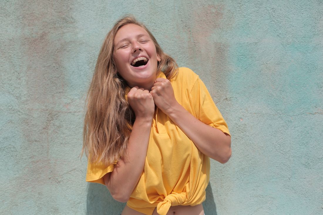 Laughing person in a yellow shirt against a teal wall, hands clasped under chin.