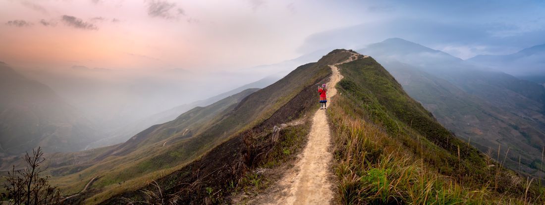 Hiker in red walking along a narrow mountain ridge at sunrise with misty valleys below