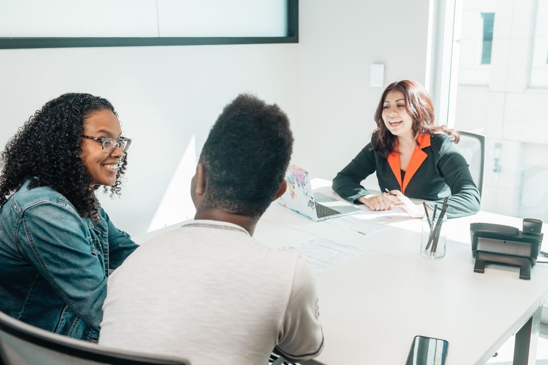 Three people in a bright conference room talking around a table with documents and a laptop.
