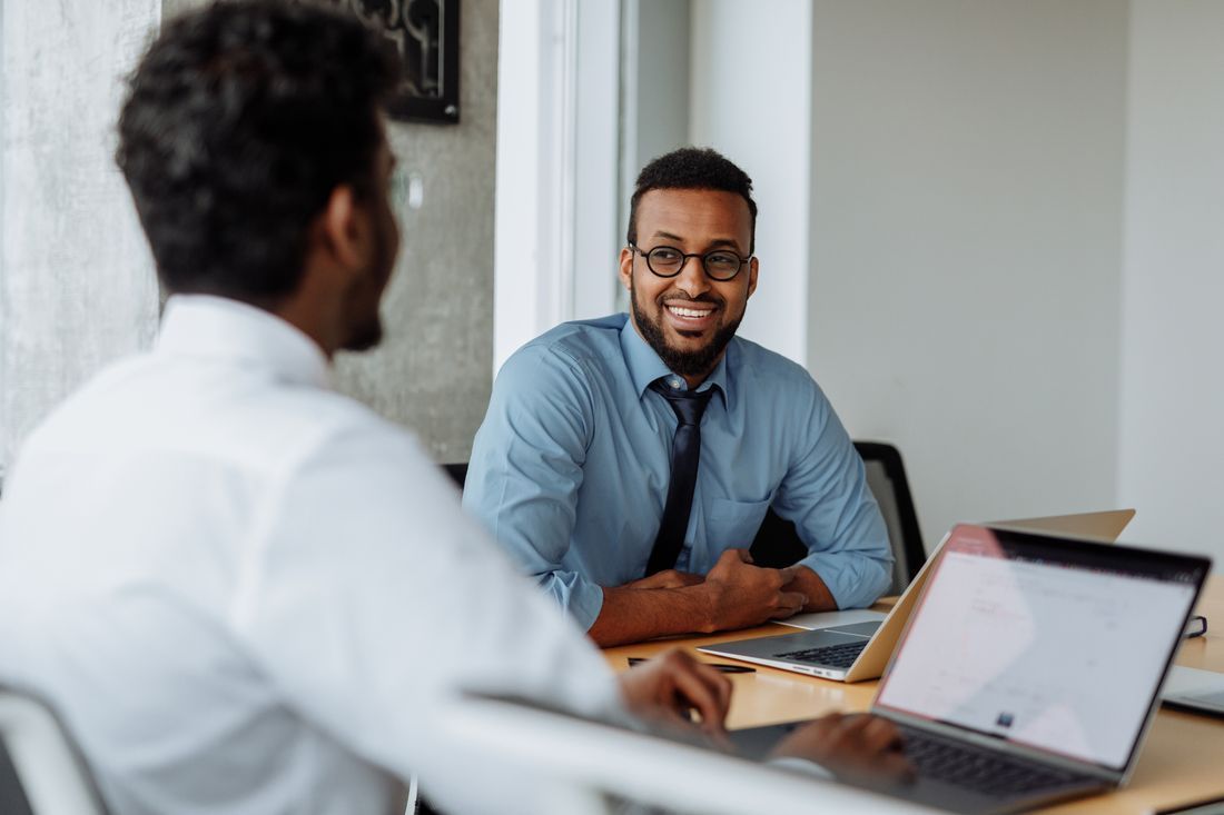 Two coworkers discussing work at a desk with laptops in a bright office