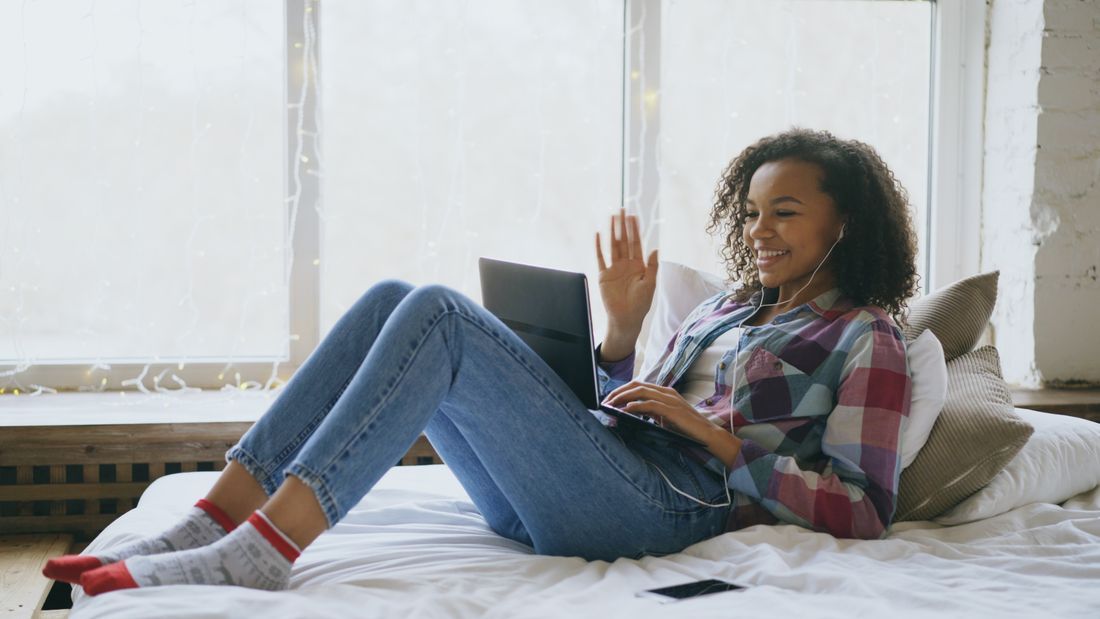 Woman reclining on a bed, smiling and waving while using a laptop in a bright room