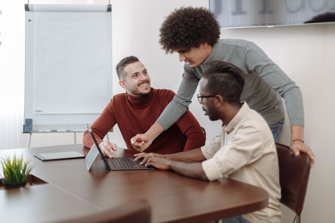 Three coworkers collaborate around a laptop at a meeting table in a bright office.