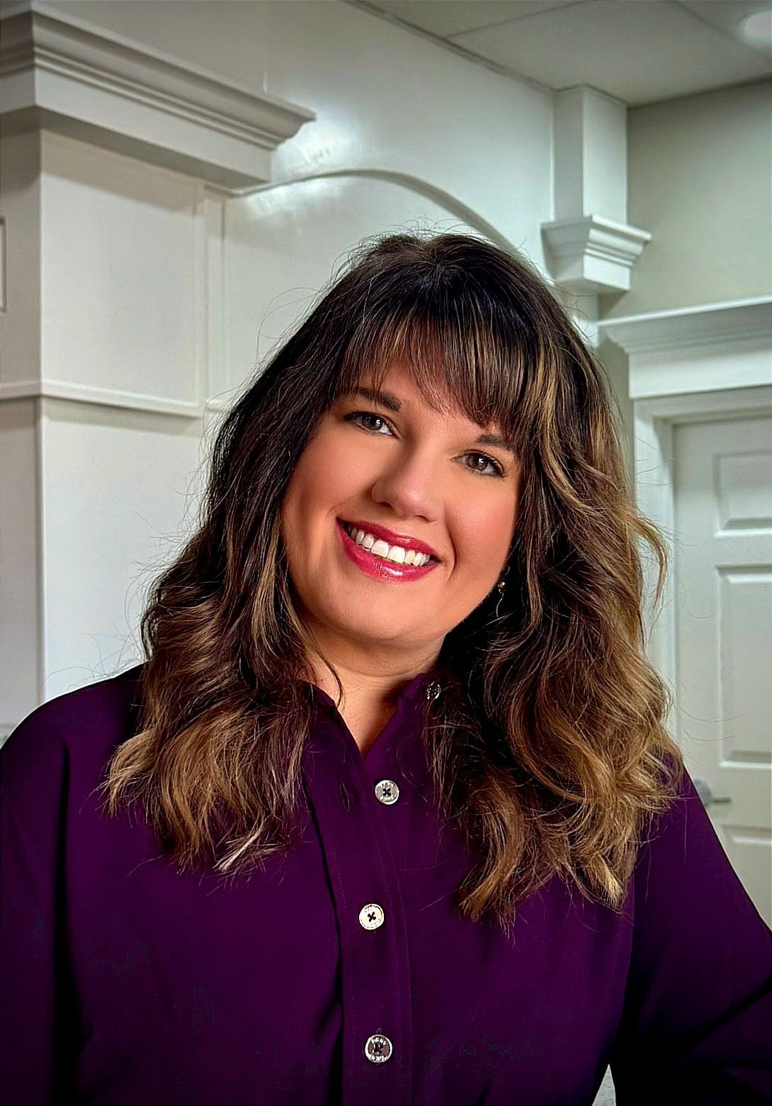 Smiling woman in a purple blouse standing indoors against a white interior background