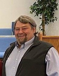 Smiling man in a light blue shirt and dark vest standing indoors near a potted tree.