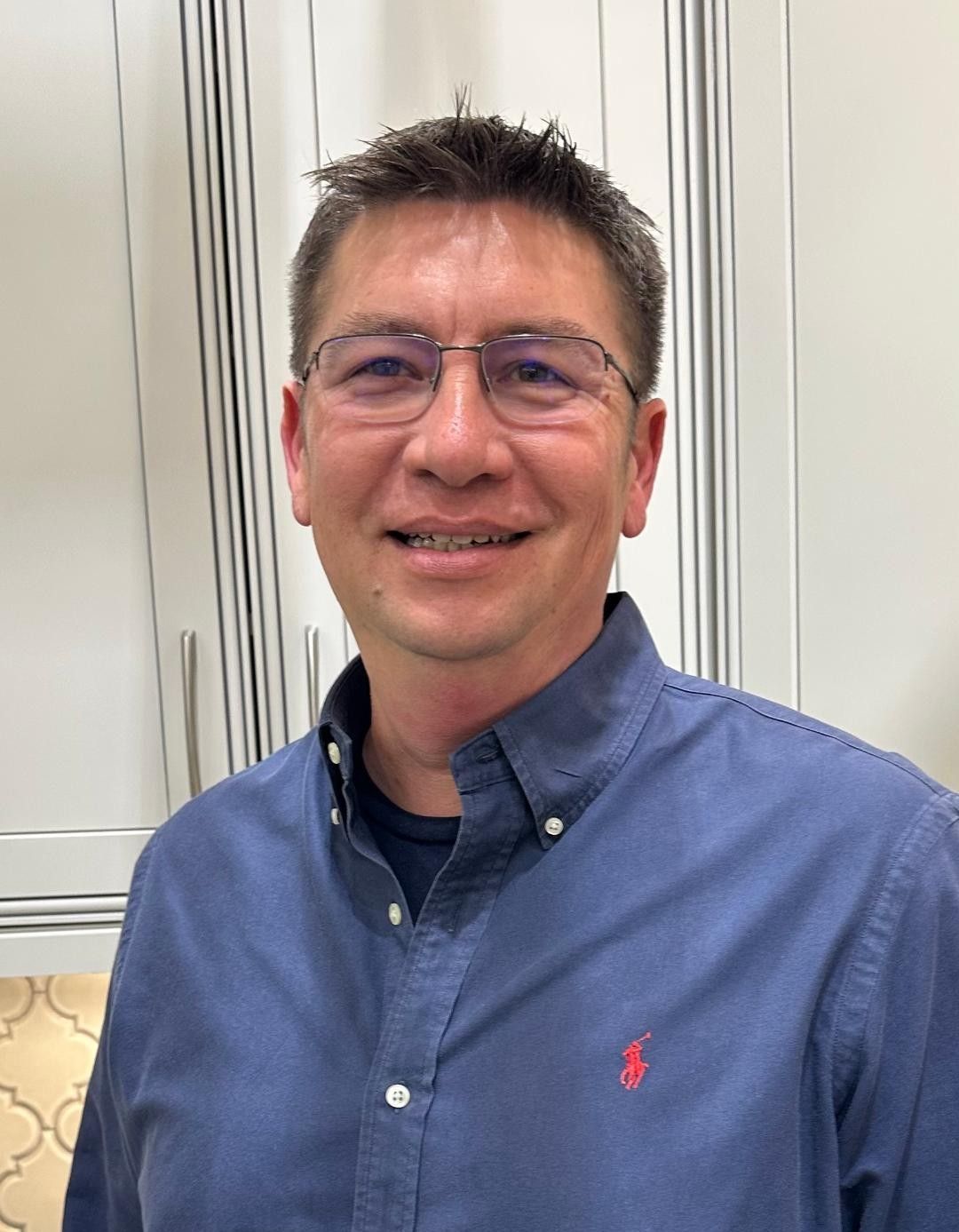 Smiling person in glasses and blue button-down shirt standing indoors against white cabinets