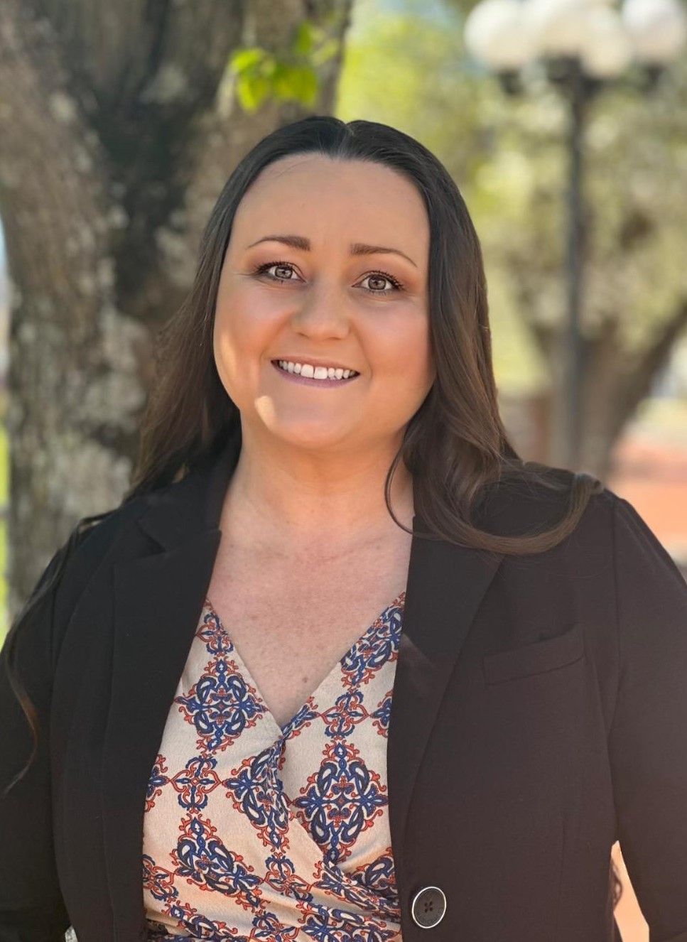 Smiling woman in a black blazer and patterned blouse standing outdoors near a tree
