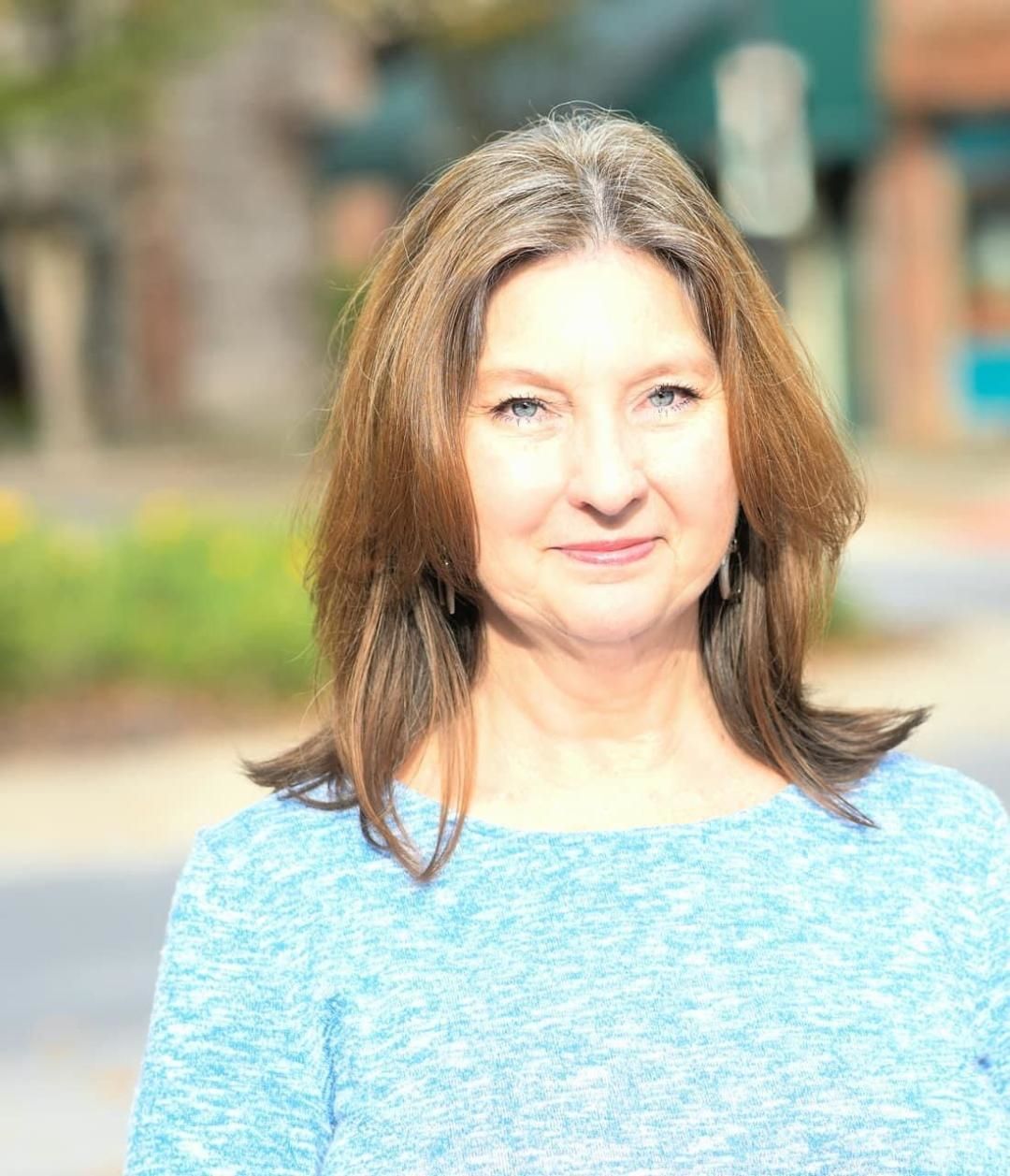 Portrait of a woman outdoors in a light blue top, standing against a softly blurred background.