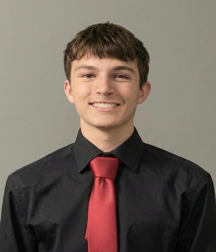 Smiling person in a black shirt and red tie against a plain gray background