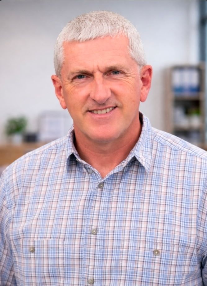 Portrait of a smiling man in a light plaid shirt, standing indoors with a blurred office background