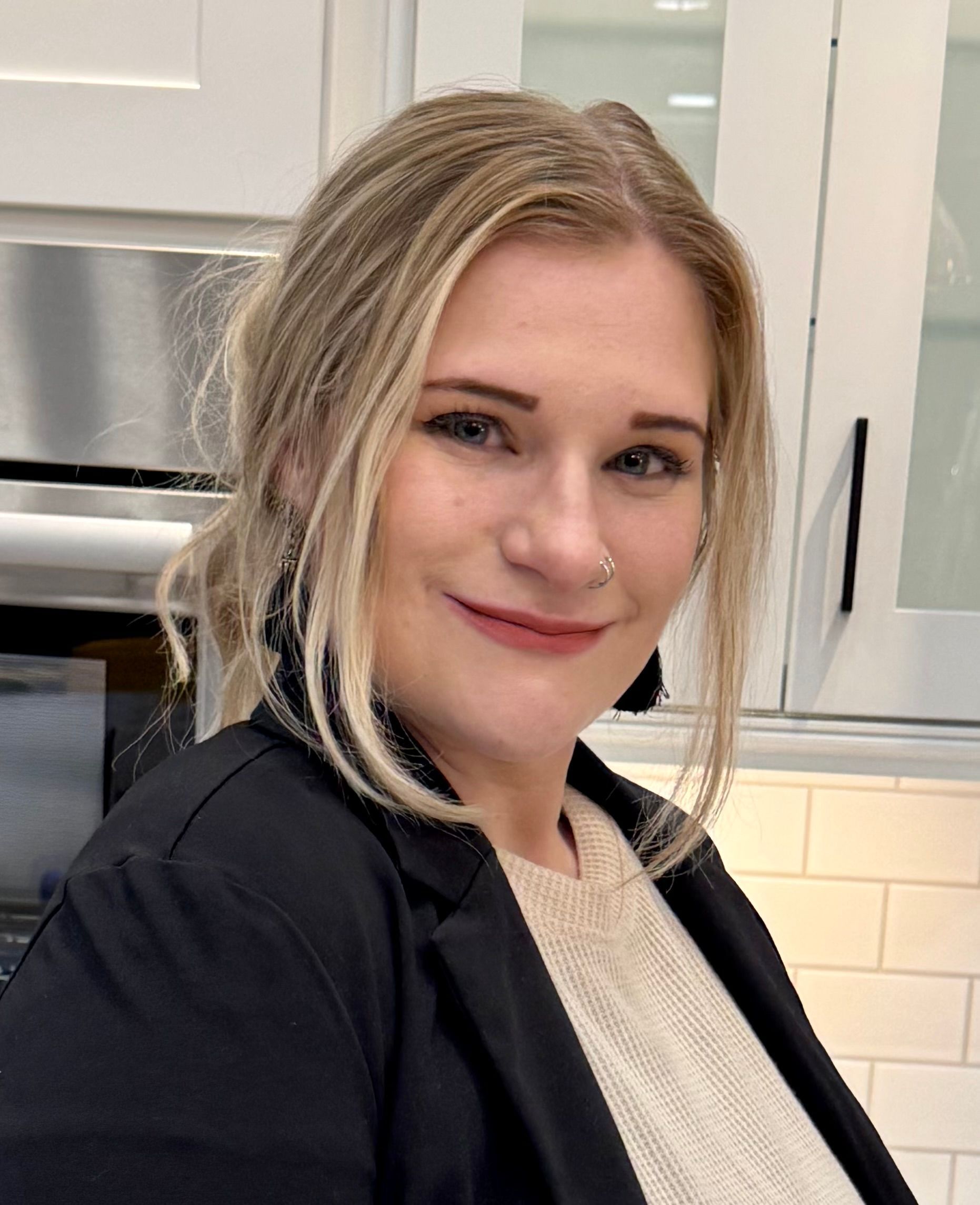 Person in a black jacket smiling in a bright kitchen with white cabinets.