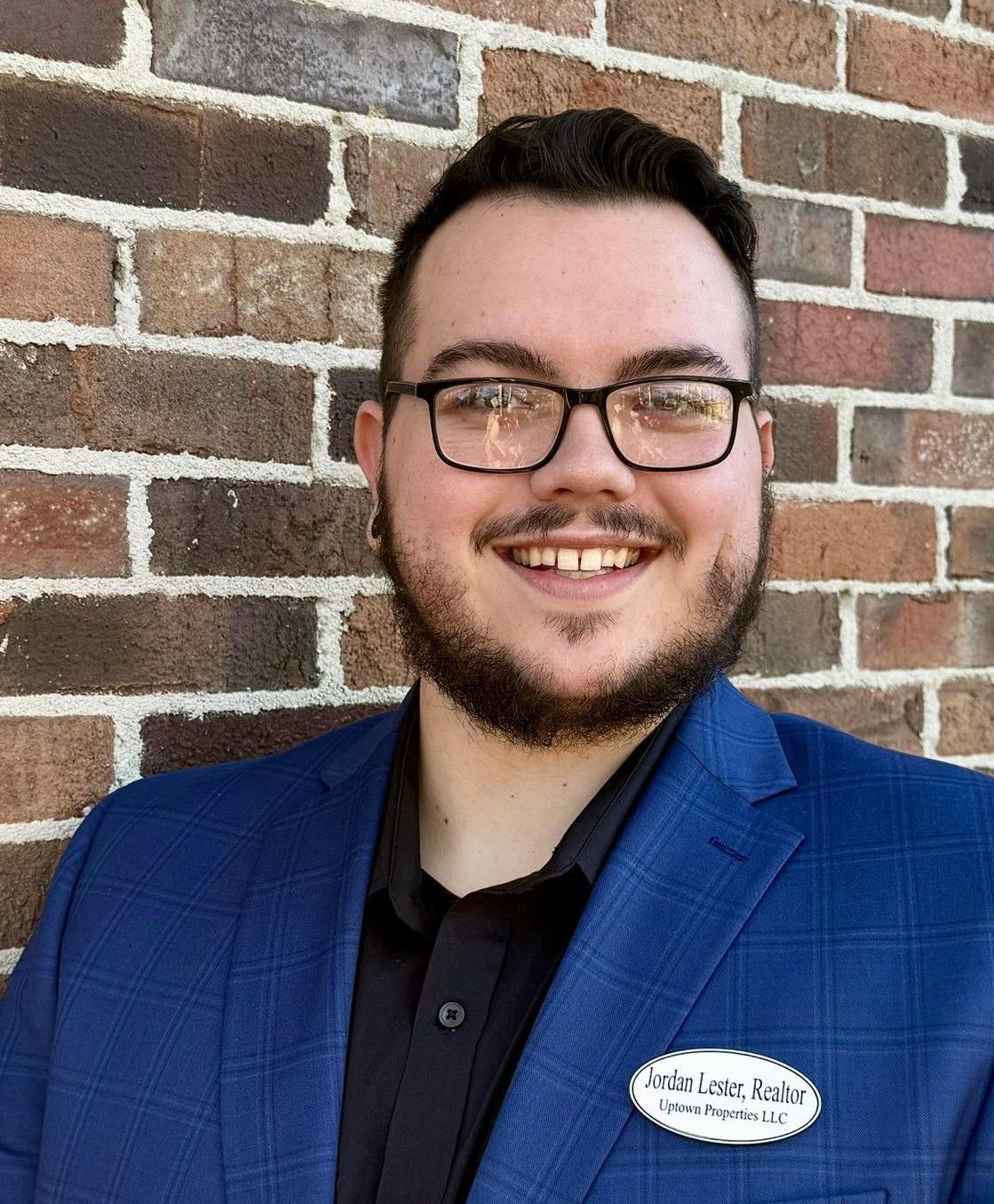 Smiling person in a blue blazer and glasses standing against a brick wall