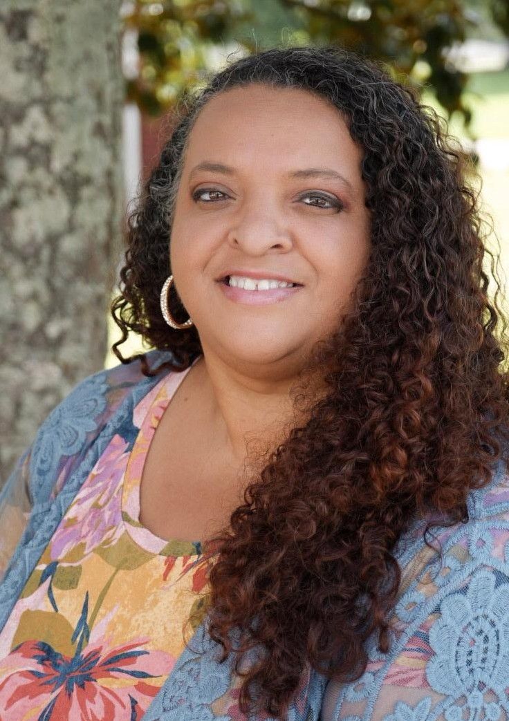 Smiling woman with curly hair in a floral top outdoors near trees and a stone wall