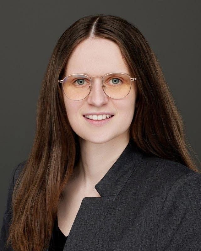Portrait of a person with long brown hair, glasses, and a dark blazer against a gray background