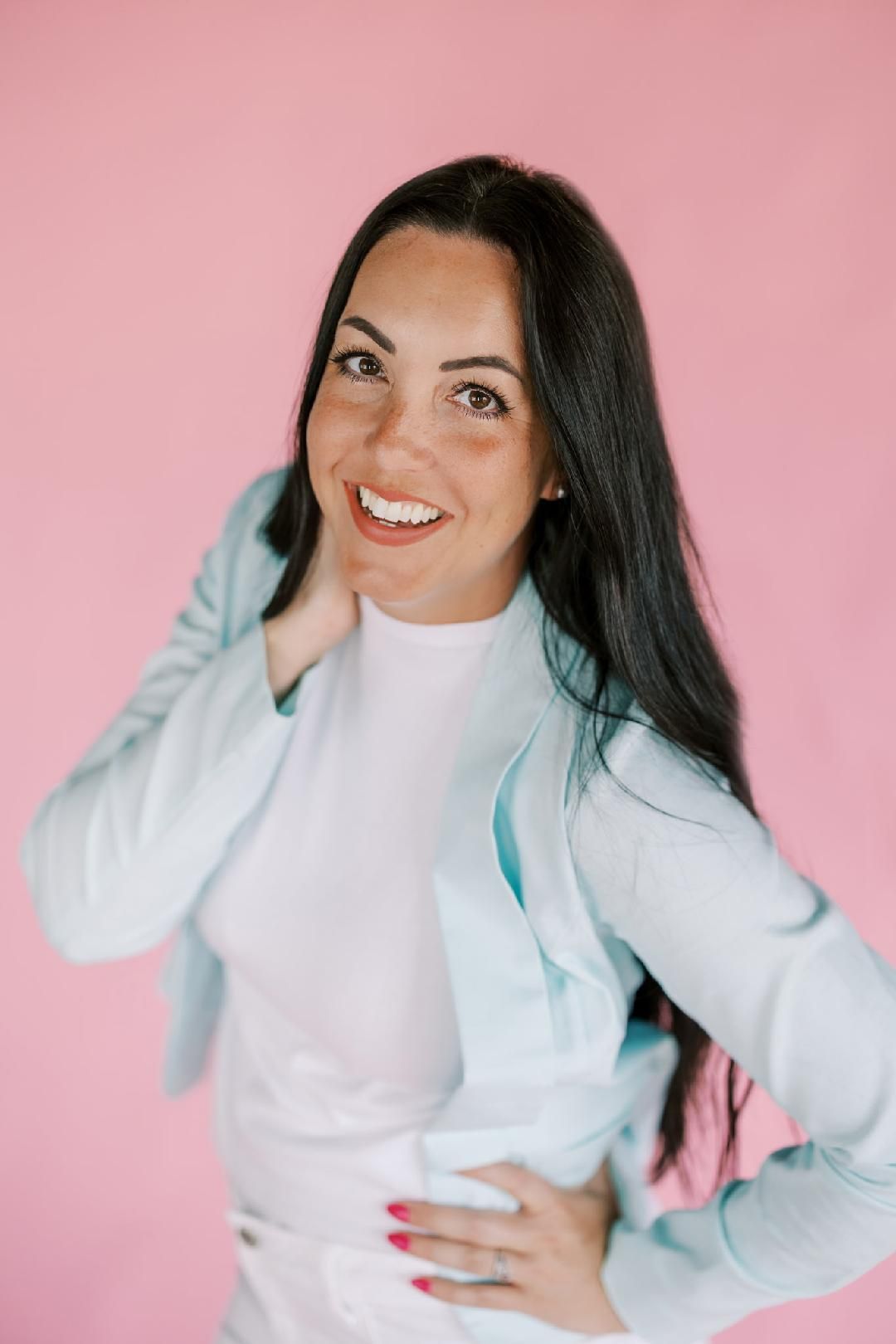 Smiling woman in white outfit with light blue jacket posing against a pink background