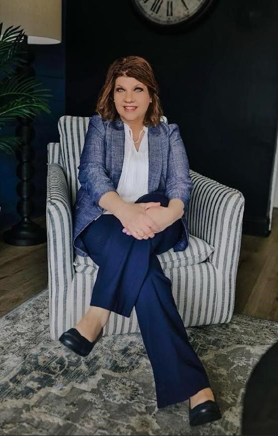 Woman seated in a striped armchair, smiling in a blue blazer and pants in a dimly lit room.