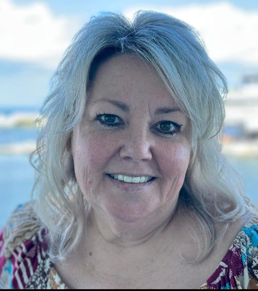 Smiling woman with blonde hair in a colorful top, standing near water with a blurred waterfront background