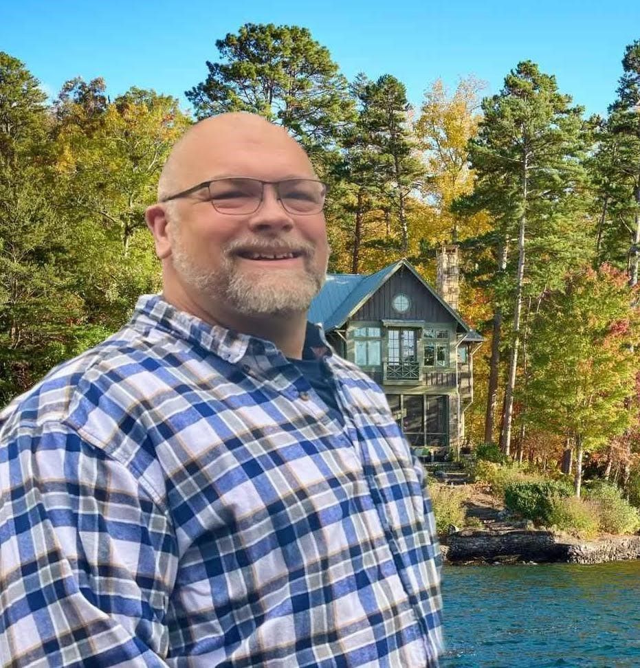 Smiling bald man in a blue plaid shirt by a lakeside cabin with trees in the background