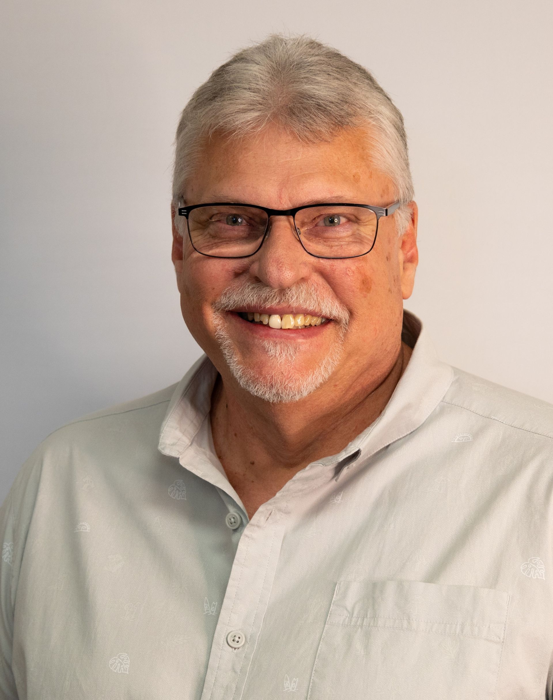 Smiling man with gray hair and glasses in a light button-up shirt against a plain background