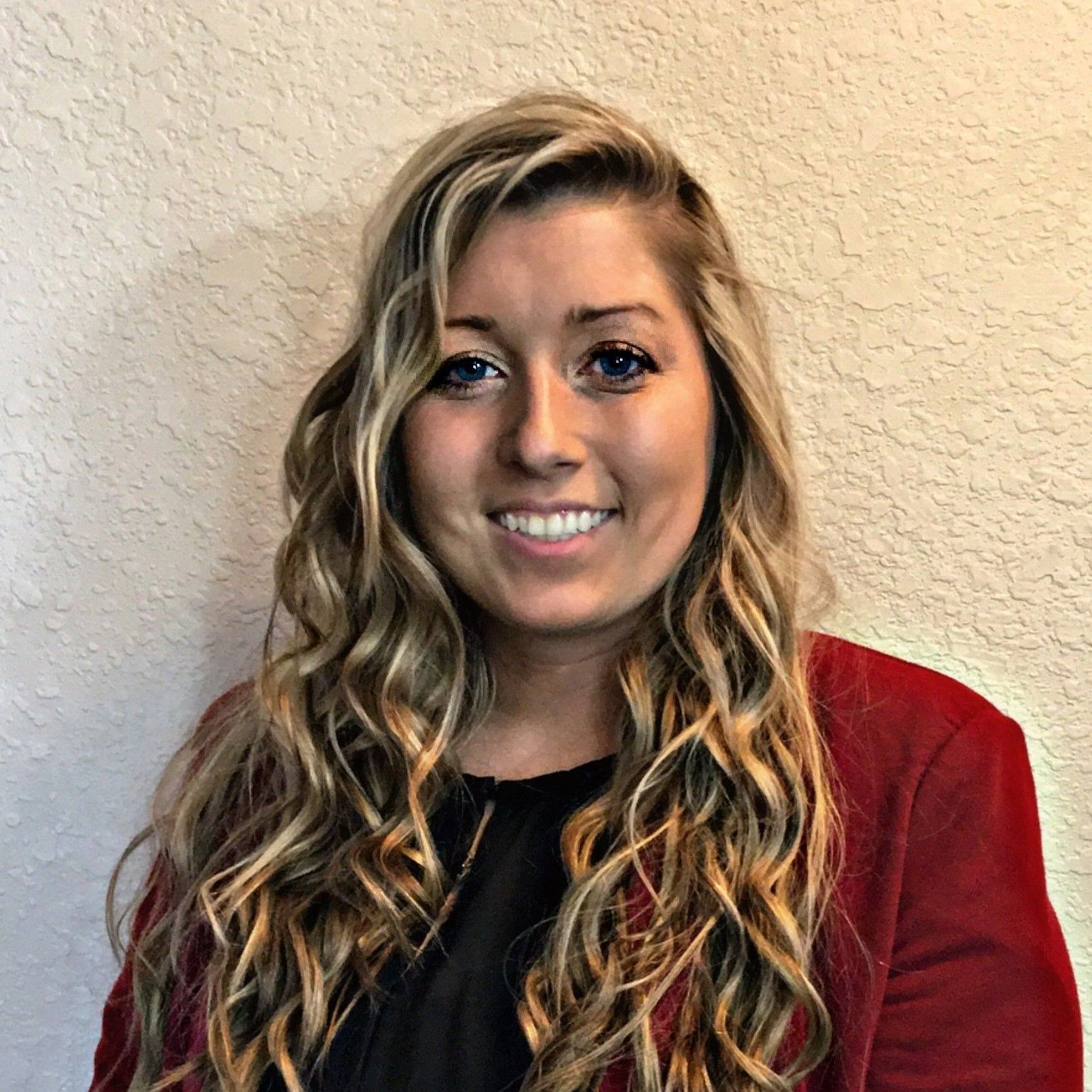 Smiling woman with long wavy blonde hair wearing a red blazer against a beige wall