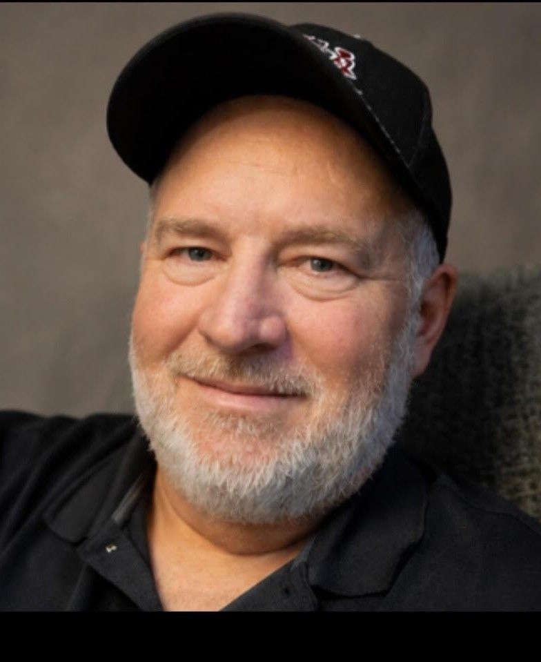 Smiling man in a black cap and shirt, with a gray beard, posing against a blurred background