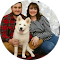 Two people sitting indoors with a white dog, all smiling and wearing casual clothes.