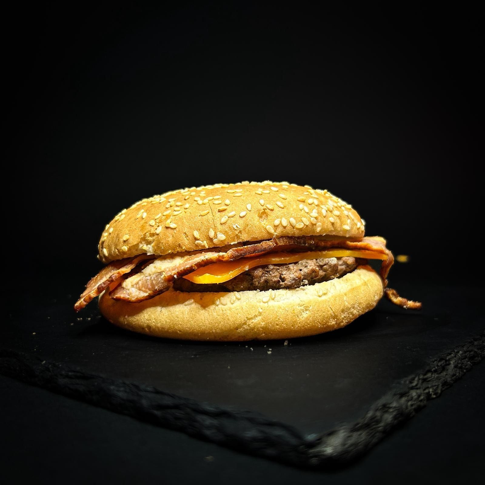 Cheeseburger al sesamo con bacon su una superficie di ardesia scura, davanti a uno sfondo nero