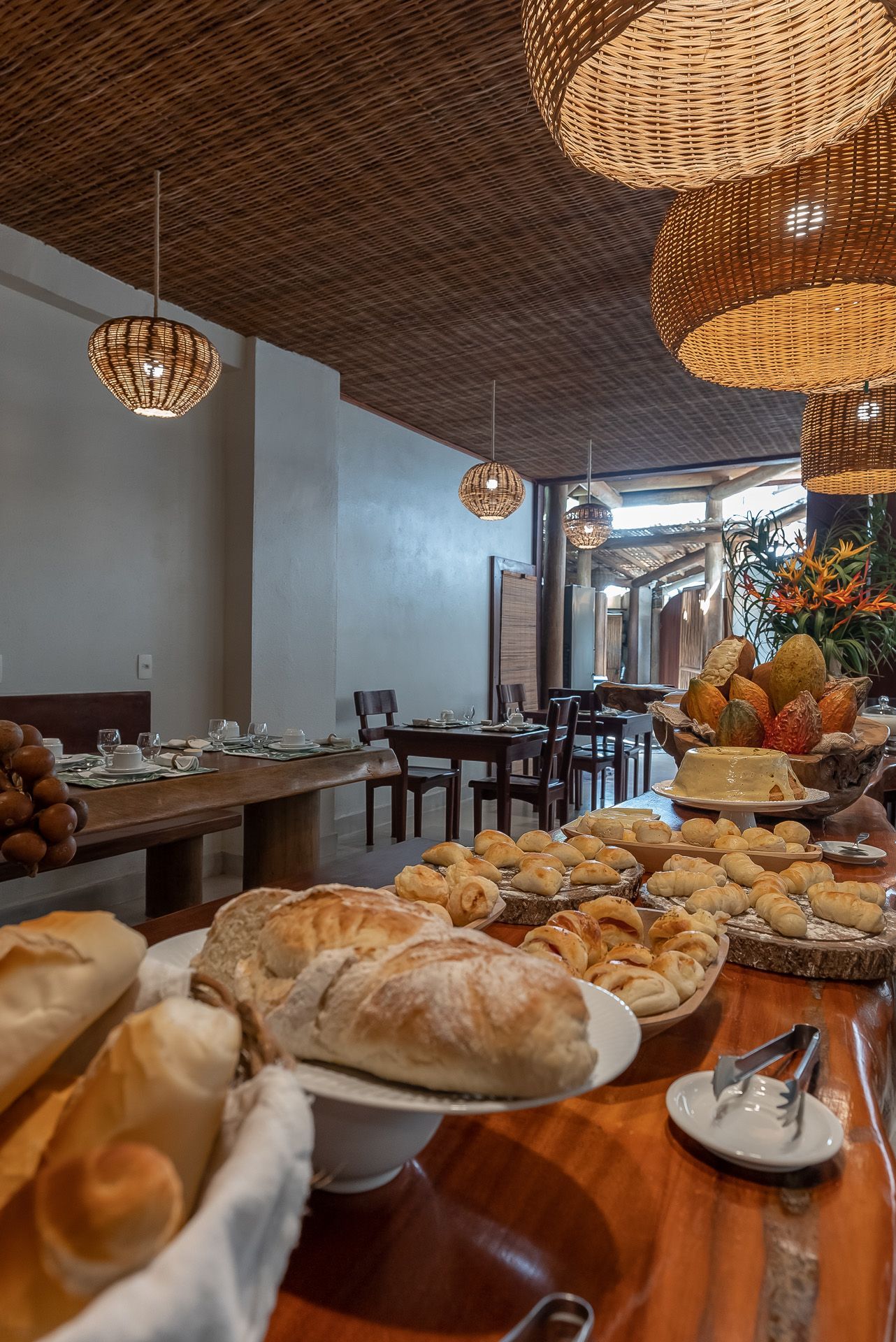 Buffet de café da manhã: pães, doces, frutas e muito mais em uma mesa de madeira. Luminárias de vime pendem de um teto trançado.