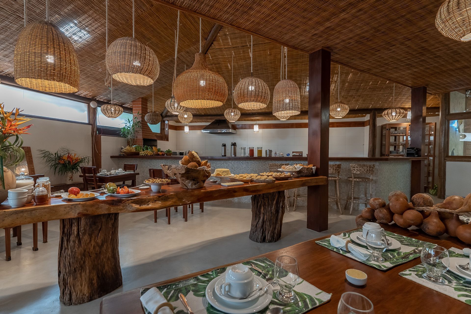 Buffet de café da manhã com mesas de madeira, luminárias pendentes e variedade de alimentos. Decoração tropical no restaurante.