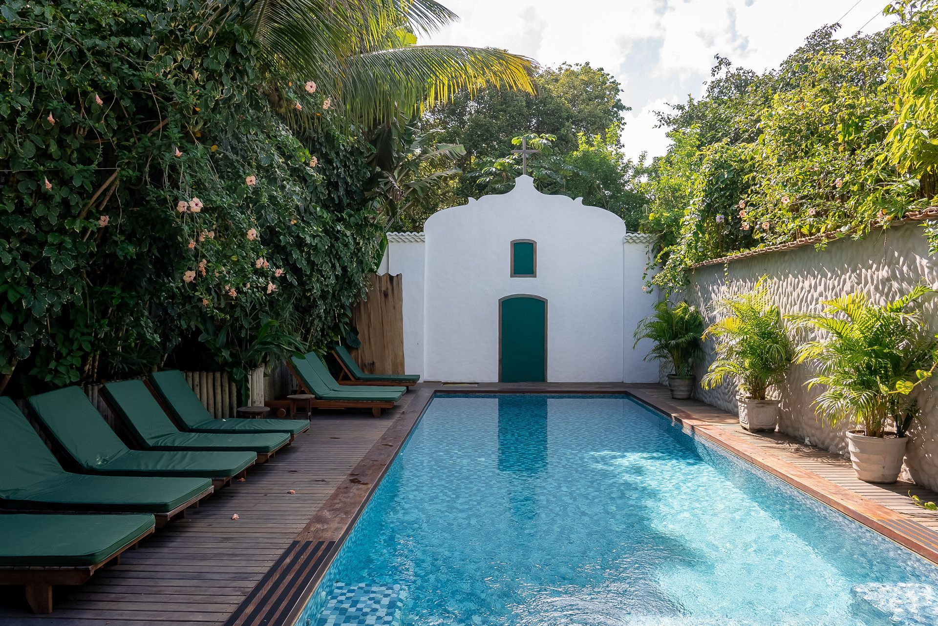 Piscina rodeada por espreguiçadeiras verdes e vegetação exuberante, com um edifício branco e uma porta verde ao fundo.
