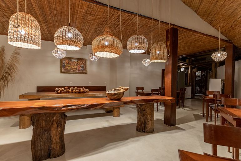 Interior de restaurante com mesas de madeira, luminárias pendentes e teto de palha.