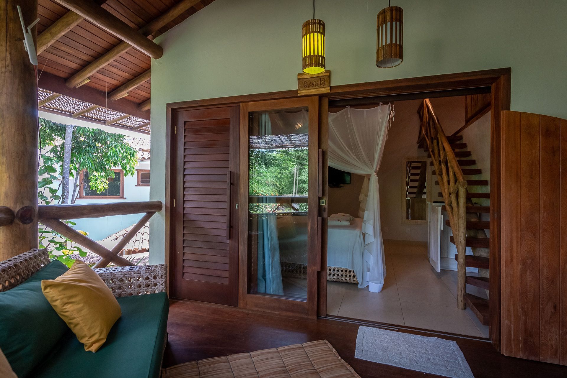 Varanda de madeira com sofá, que dá acesso a um quarto com cama e escada. Vegetação à vista.
