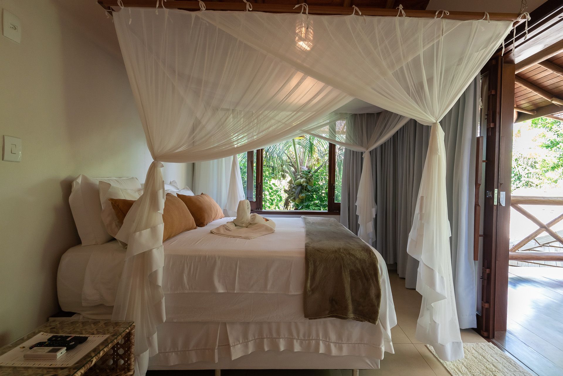 Quarto com cama de dossel, roupa de cama branca, portas abertas para a varanda e janela com vista para o verde.
