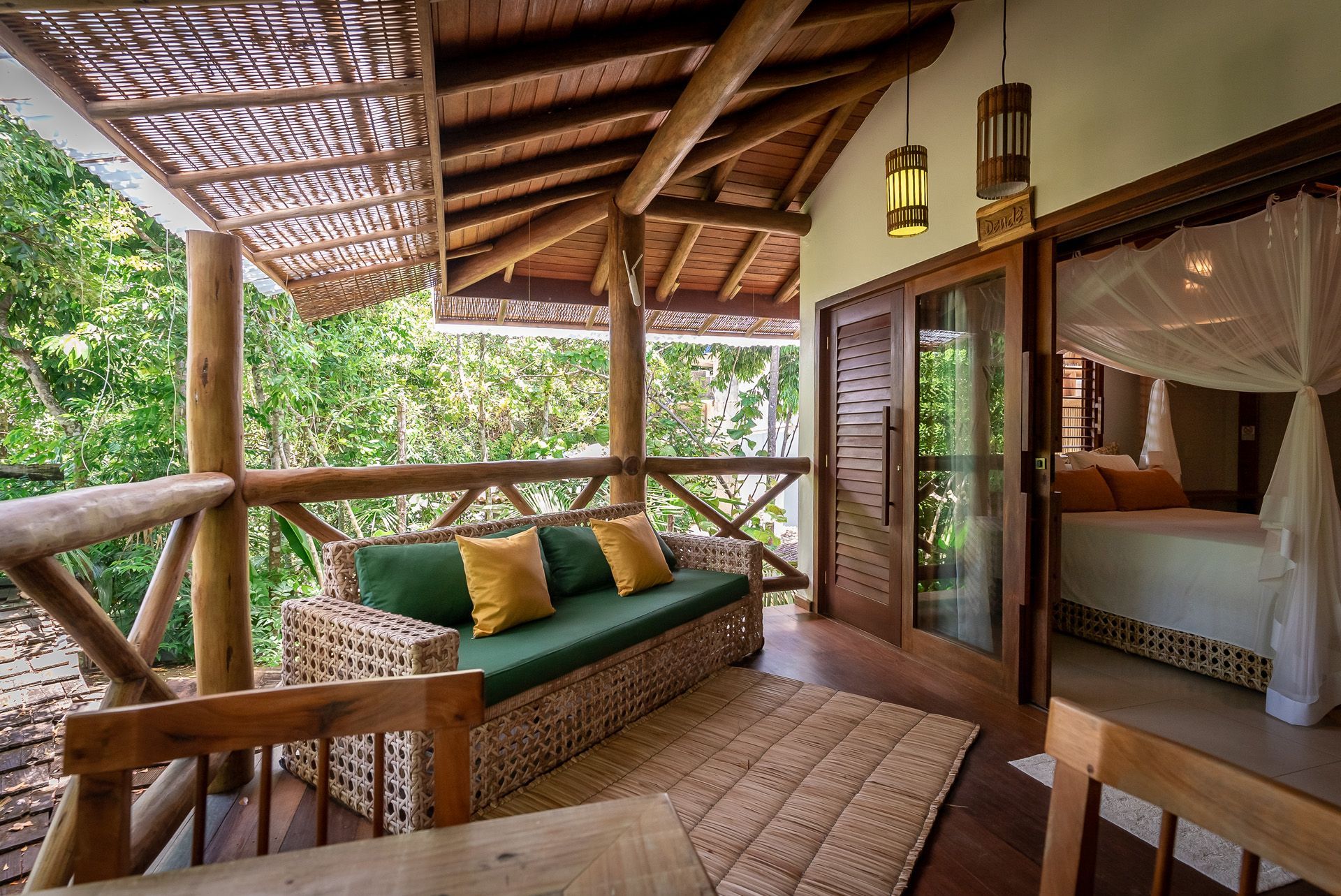 Varanda com sofá de vime, cama visível através de portas abertas, floresta ao fundo.