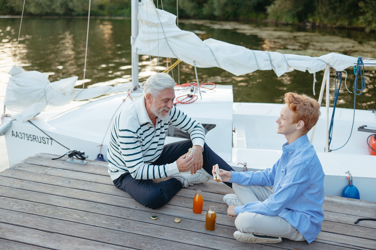 Two people sit on a dock beside a white sailboat, sharing drinks by the water.