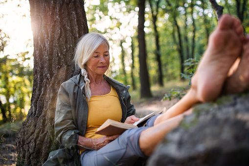 White hair old lady reading a book under the tree, relaxing legs on top of stone