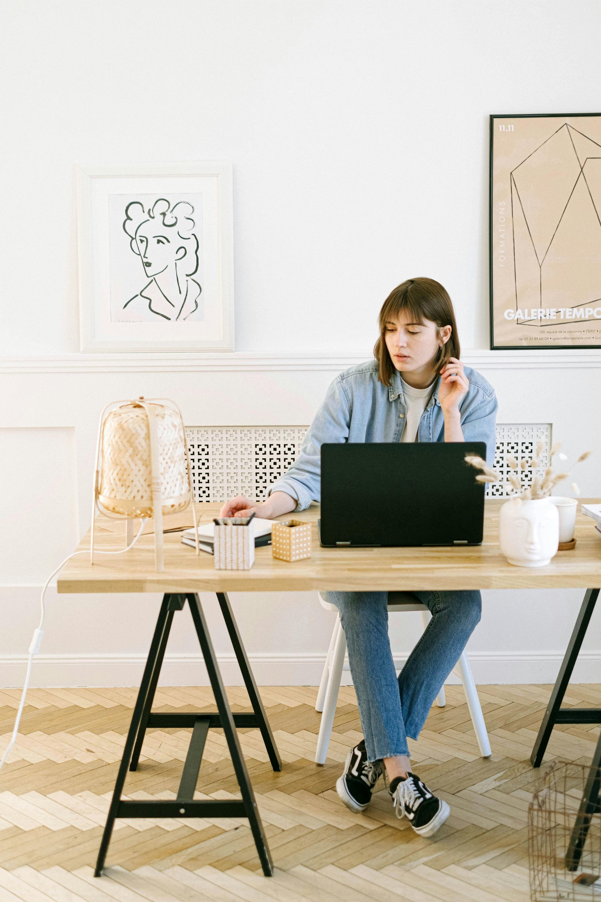 Woman in denim at a desk using a laptop, looking focused.
