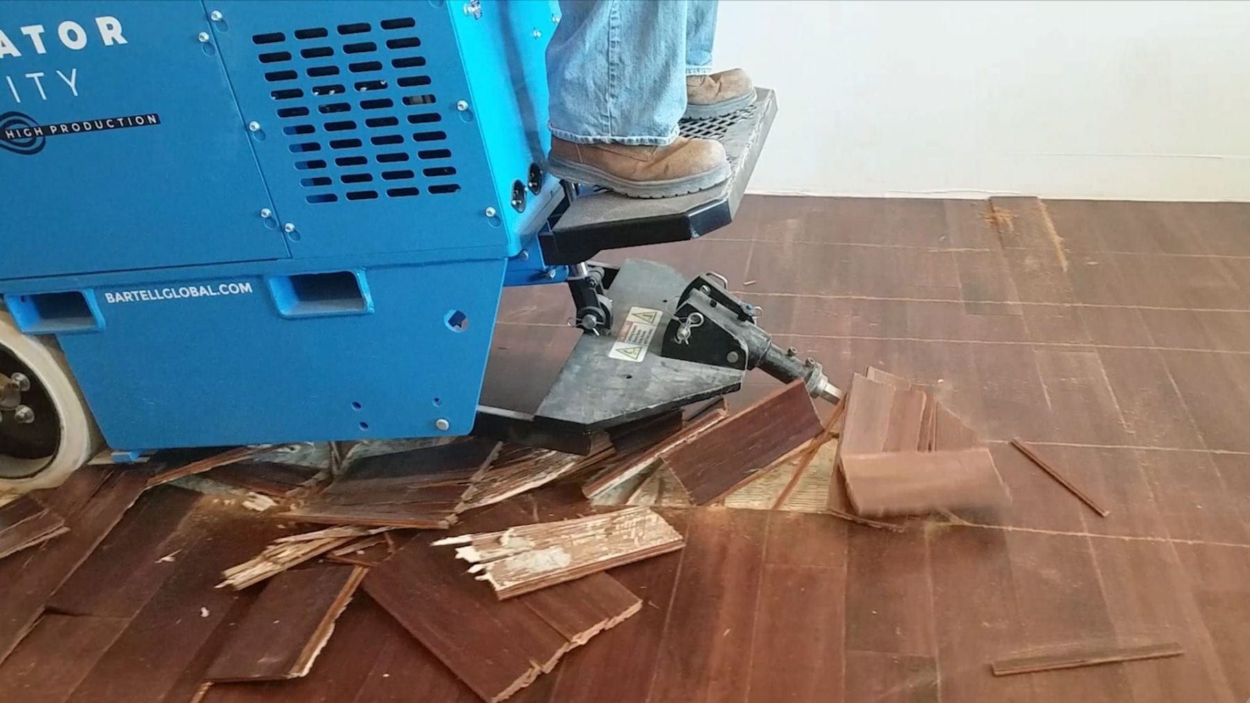 A worker using a blue floor scraper machine to remove wooden floorboards.