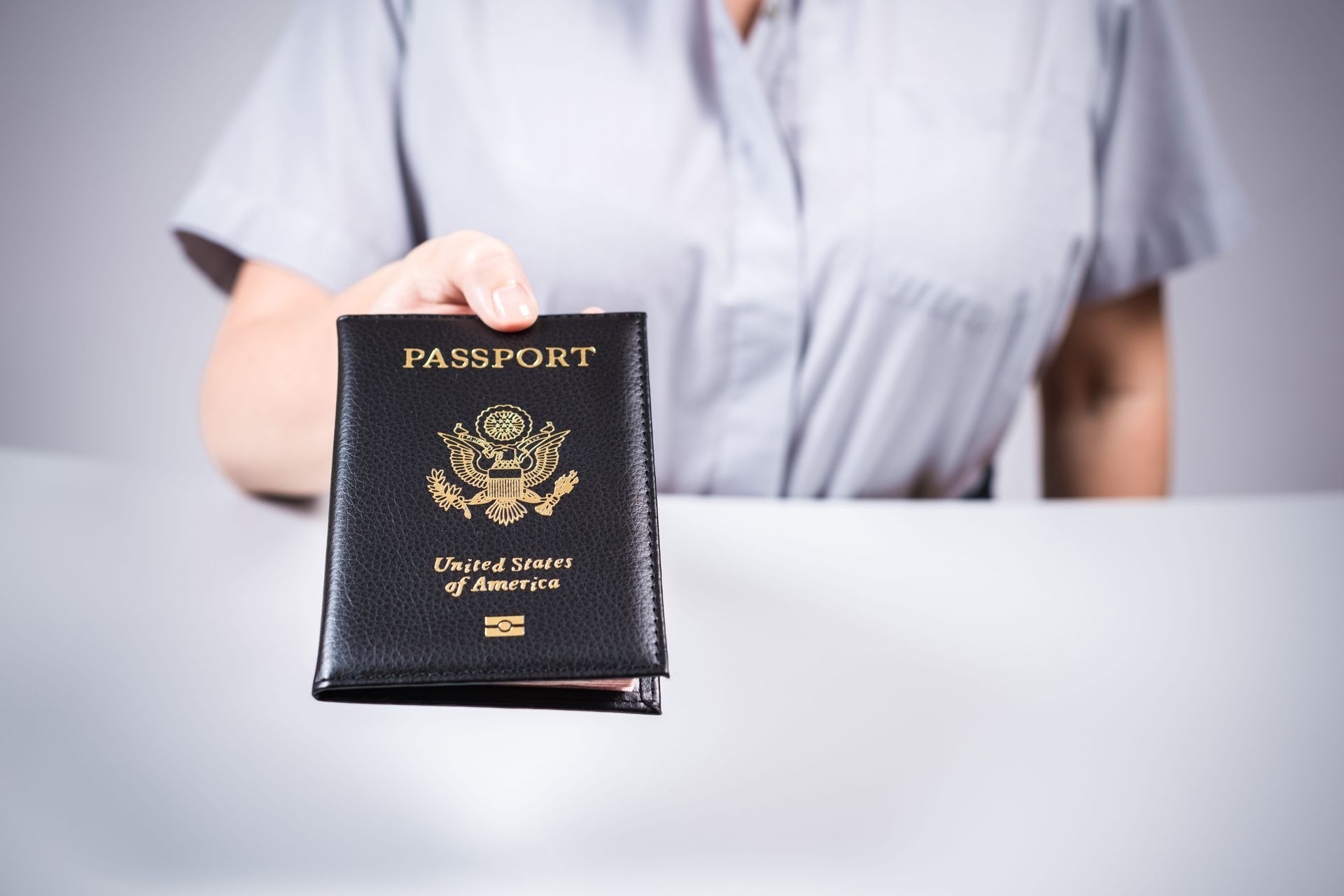 Hand holding a black passport on a white table, with a person in a light shirt in the background