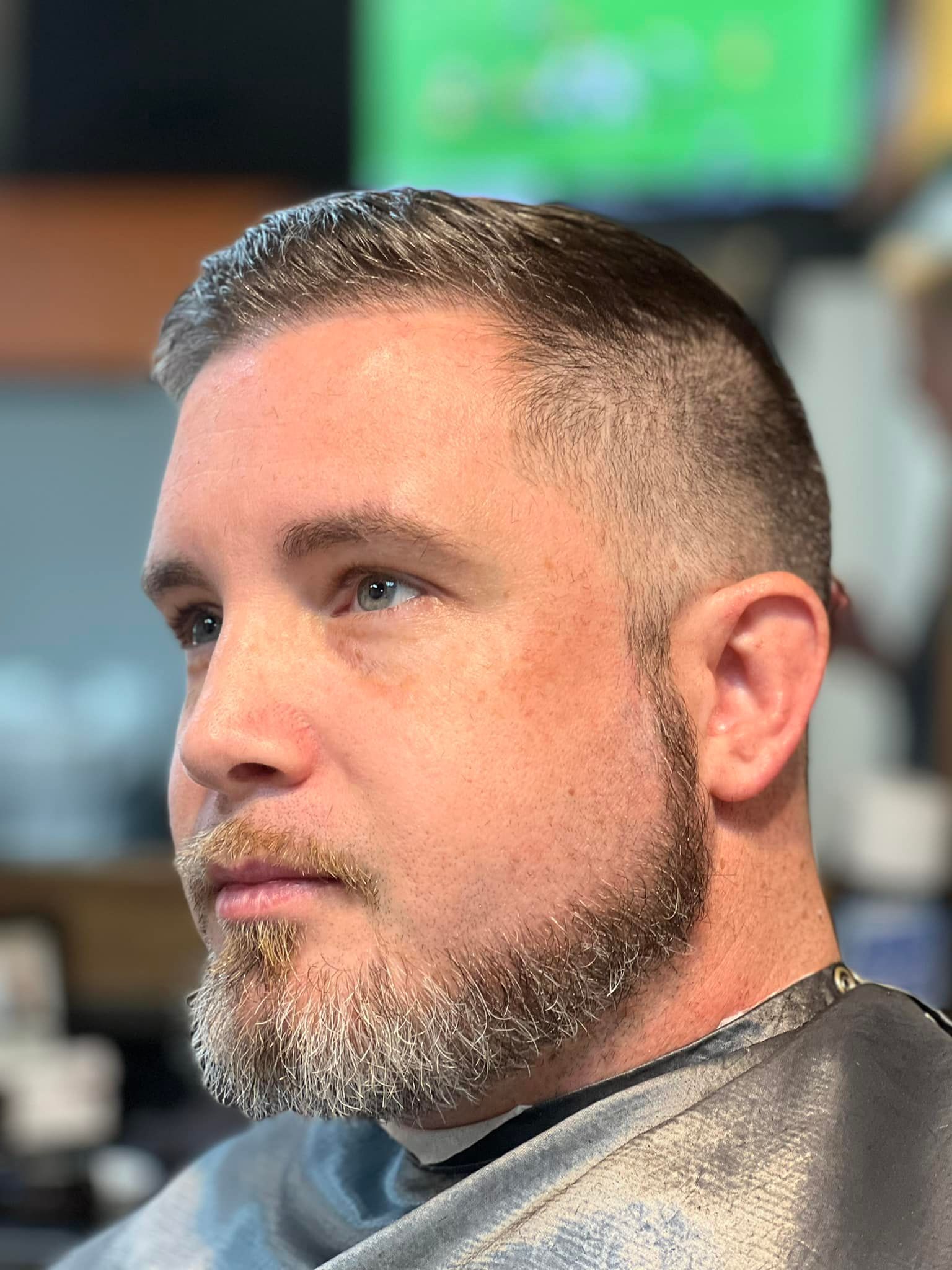 Close-up of a man with a short beard and fade haircut in a barber chair, looking to the side.
