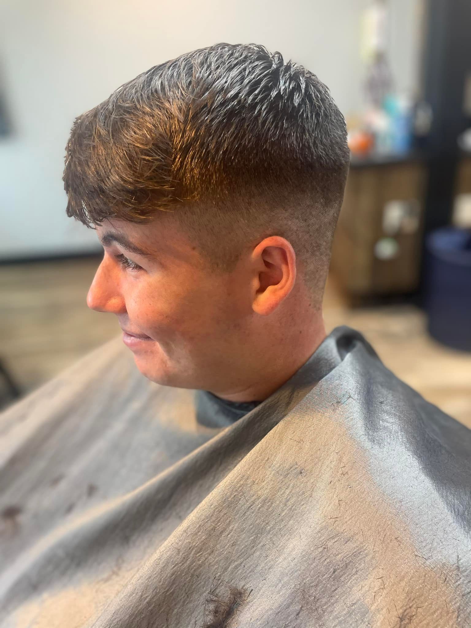 Person with a fade haircut, wearing a gray barber cape, sitting in a barbershop chair and smiling in profile.