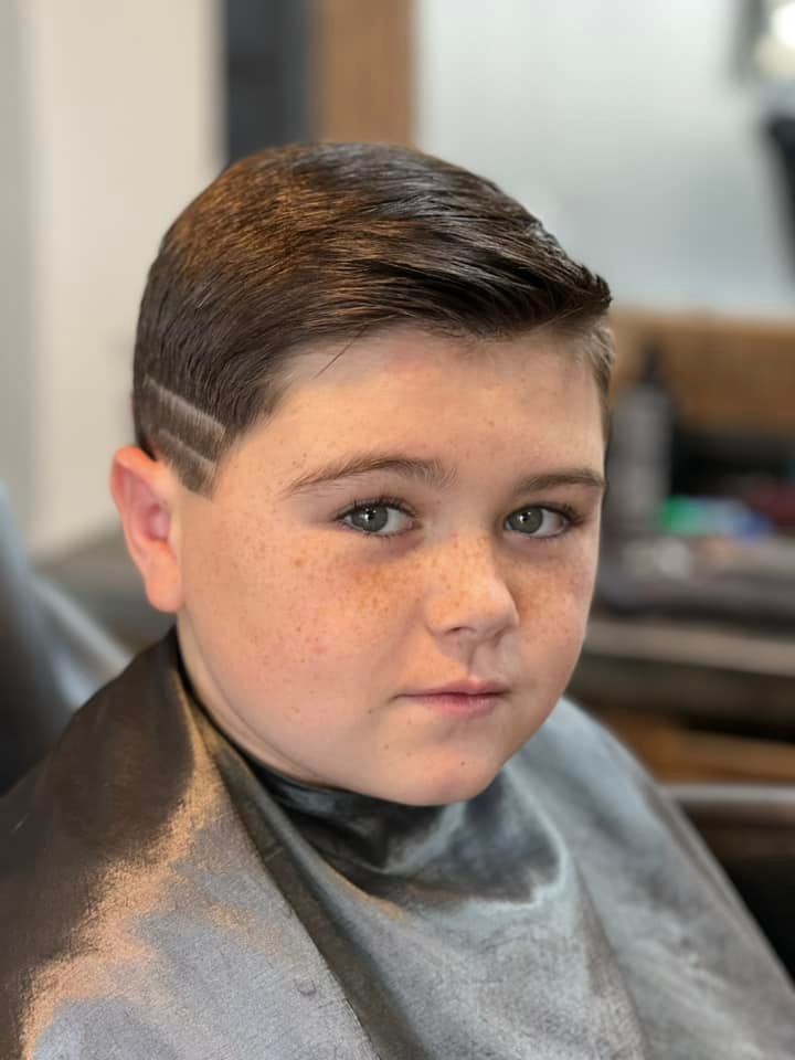 Young person with short dark hair and faded sides, wearing a gray cape in a salon chair, looking at the camera.