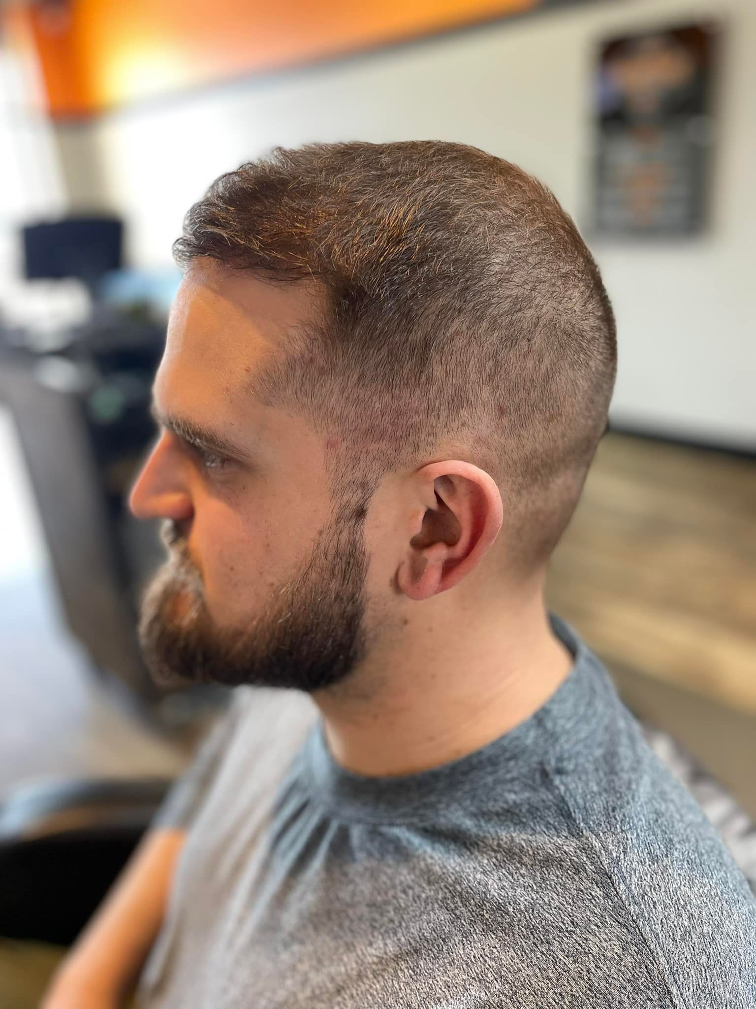 Side-profile of a bearded person with a short haircut, seated indoors in a blurred salon or barbershop.